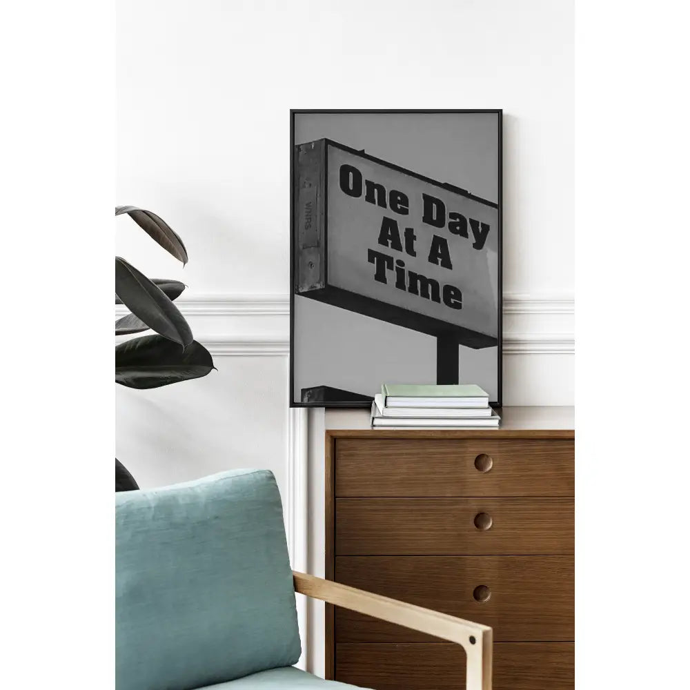 Framed artwork with 'One Day At A Time' text on a wooden dresser with a chair and books.